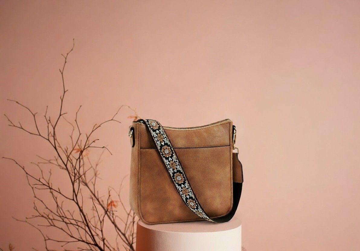 Brown minimal leather crossbody bag with floral strap on a pedestal against a soft pink background.