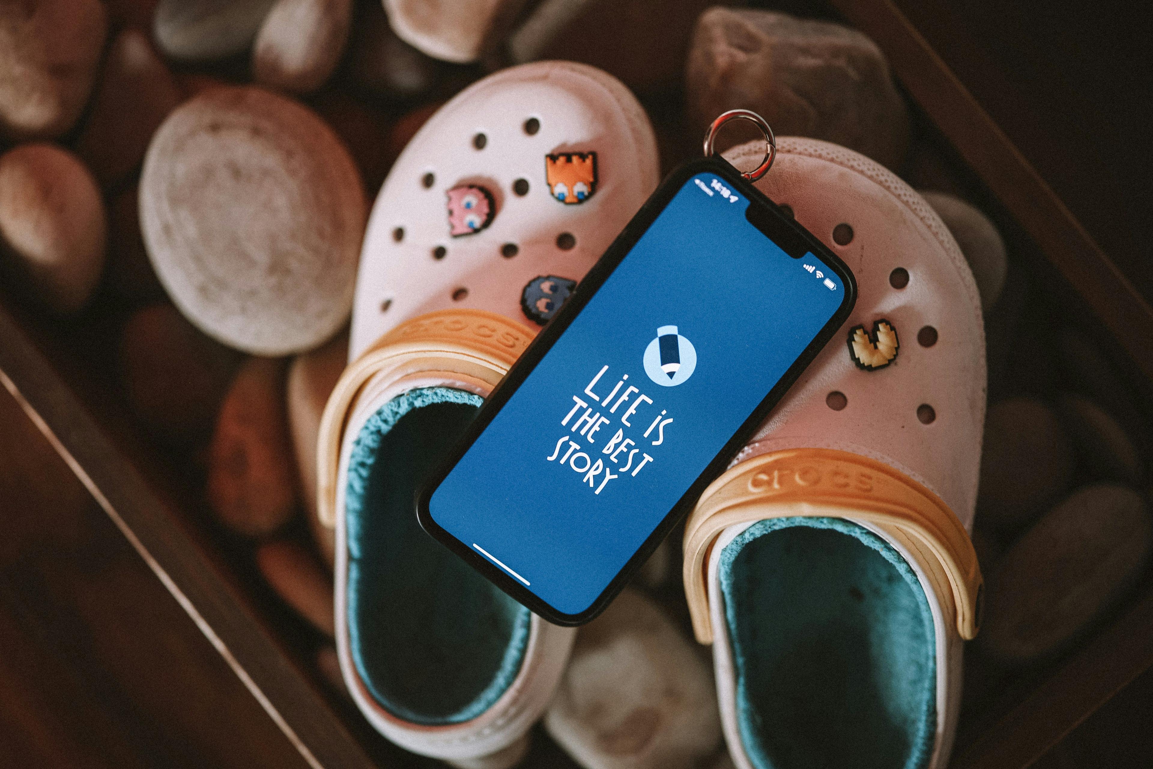 A pair of white Crocs with colorful Jibbitz charms placed on smooth stones, with a smartphone resting on top displaying the message "Life is the best story" on a blue screen.