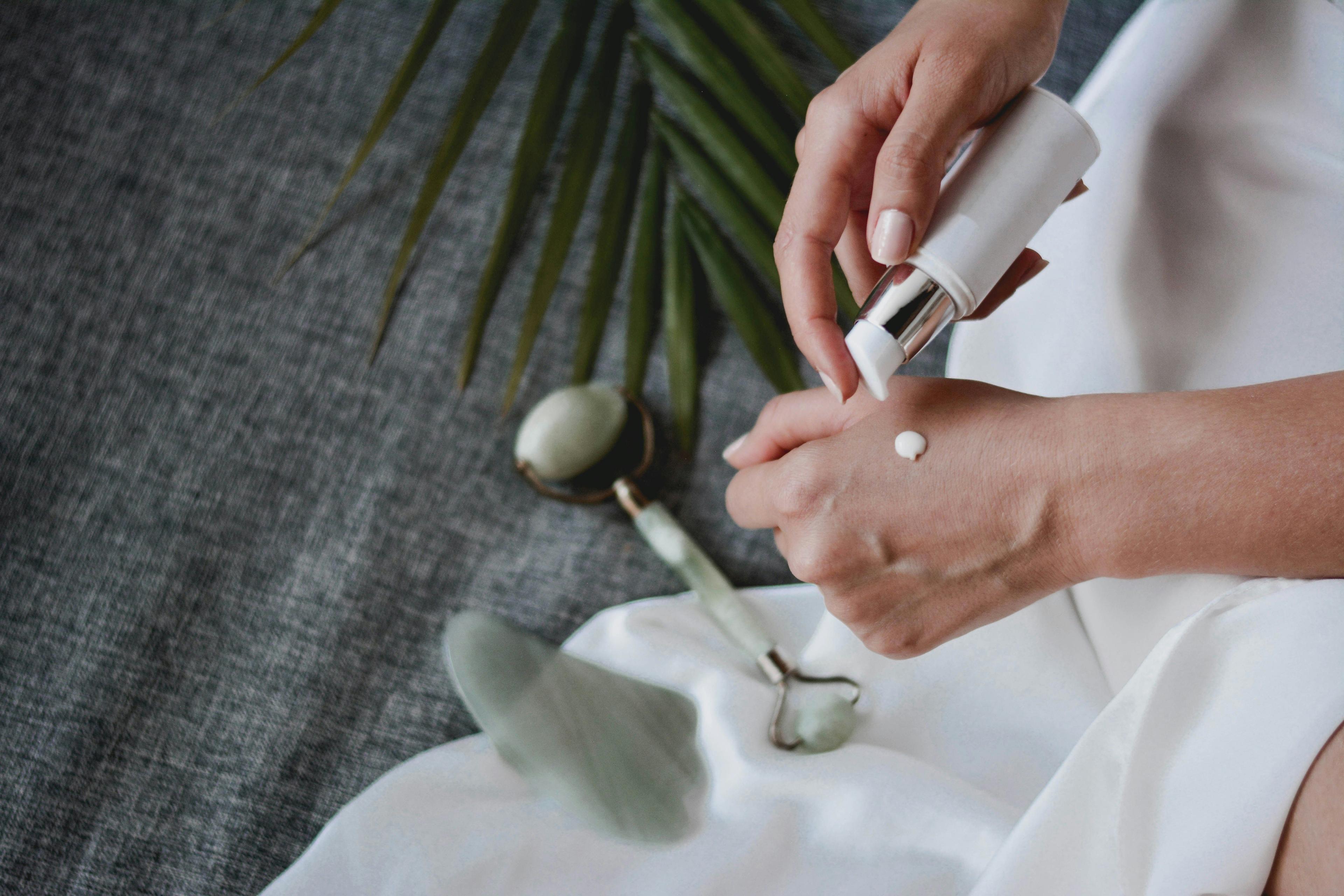 Close-up of a woman applying skincare cream to her hand with a white pump bottle, next to a jade roller and gua sha tool on fabric background.